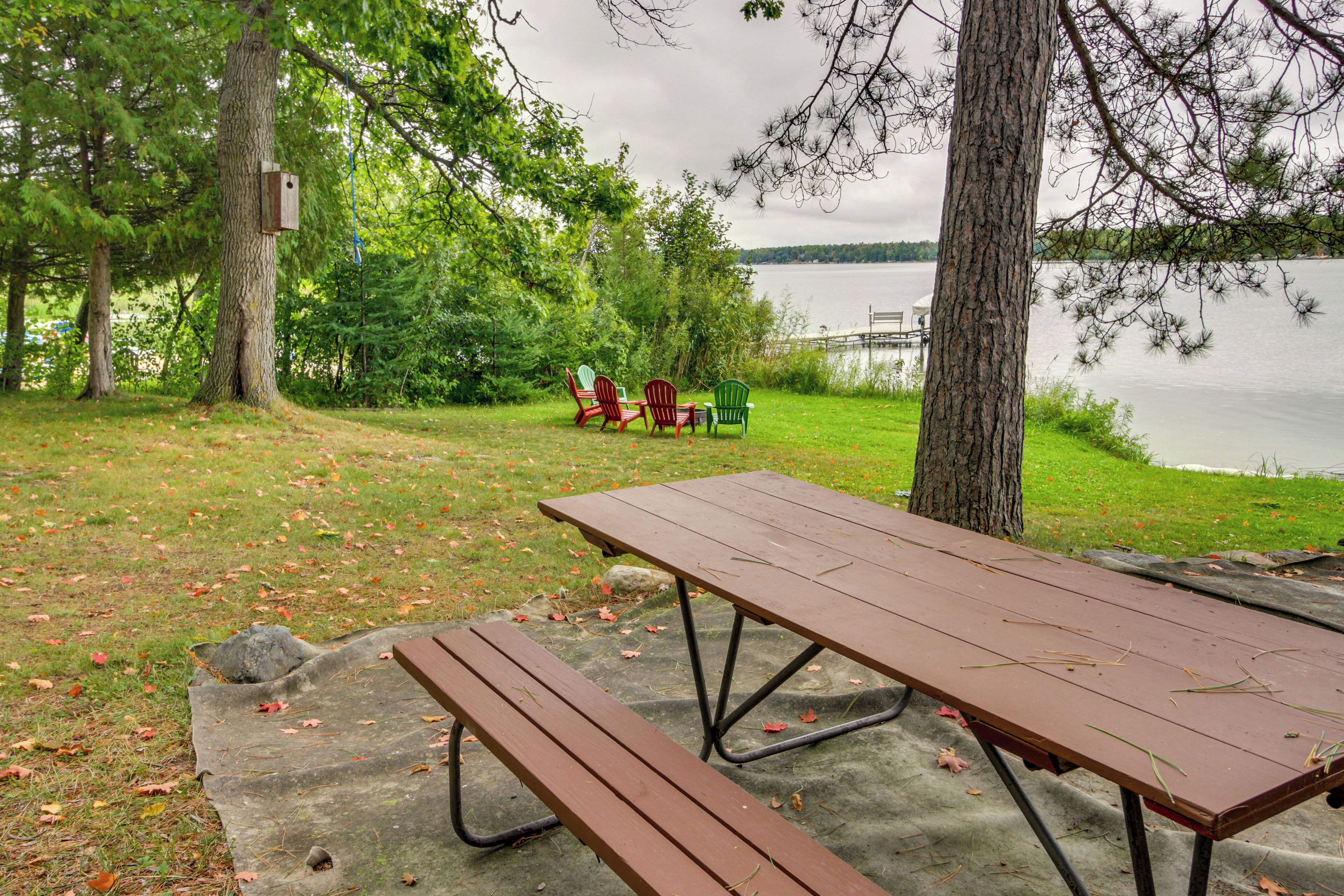 A picnic table with benches, a birdhouse, and chairs near a lake.