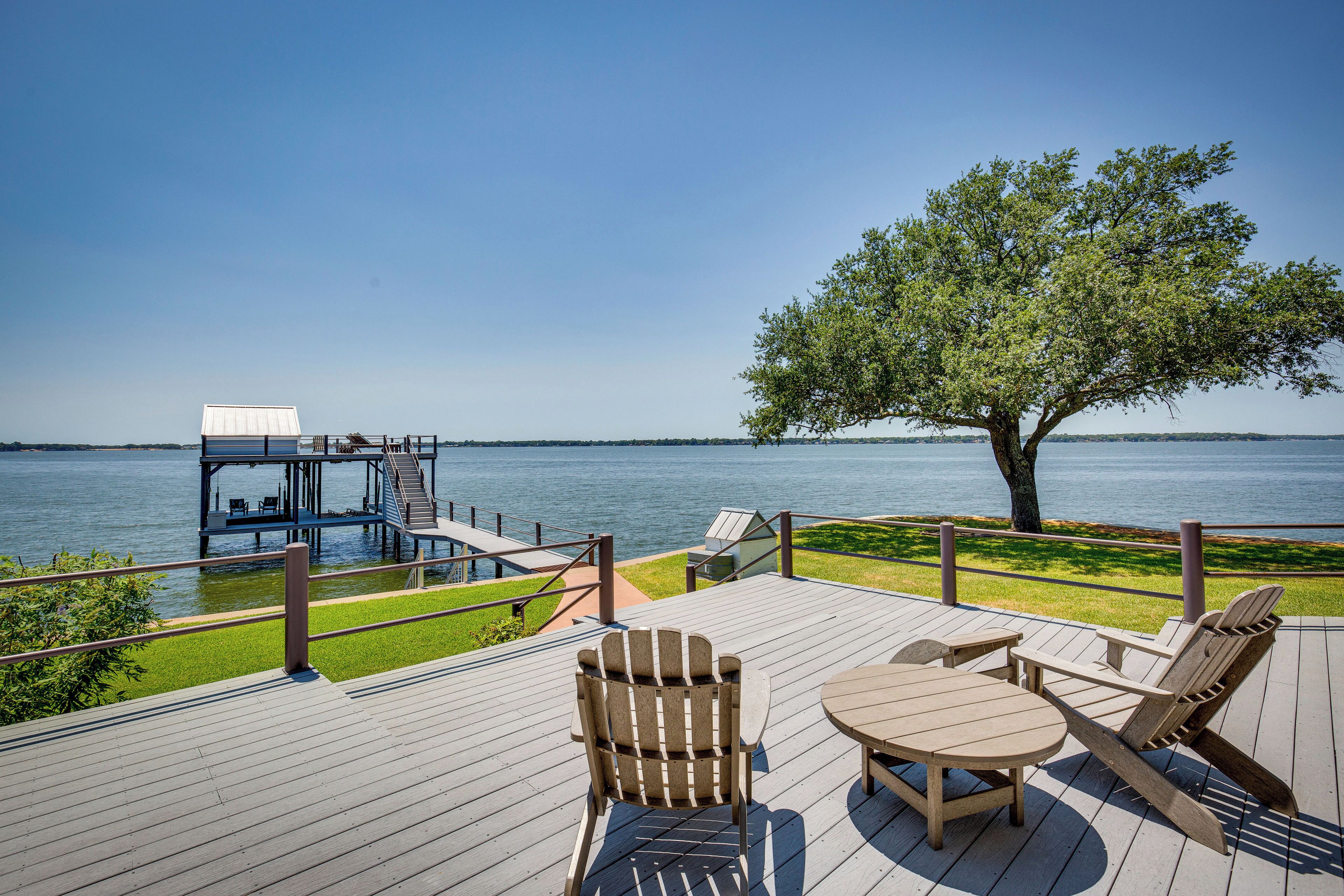 Une terrasse en bois, agréée de mobilier d’extérieur, qui offre une vue sur une étendue d’eau, un arbre et une petite construction surmontée d’un toit blanc.