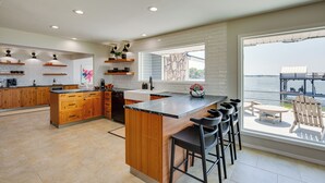A modern kitchen with wooden cabinets, a central island, and a large window overlooking a body of water. - Cedar Creek Lake Views: Gorgeous Mabank House! (Mabank)