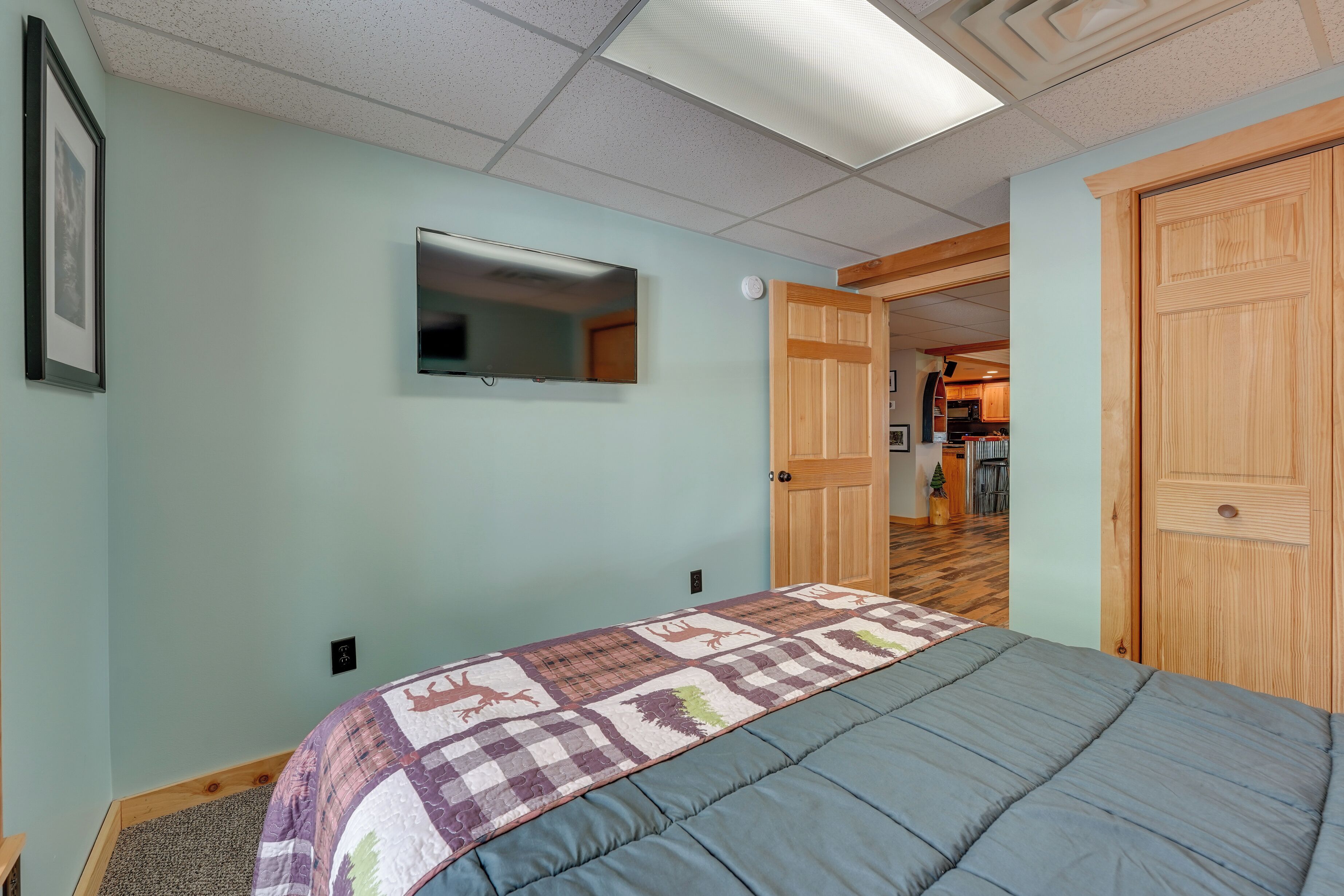 A bedroom with a bed, a wall-mounted TV, and a door leading to a kitchen area.