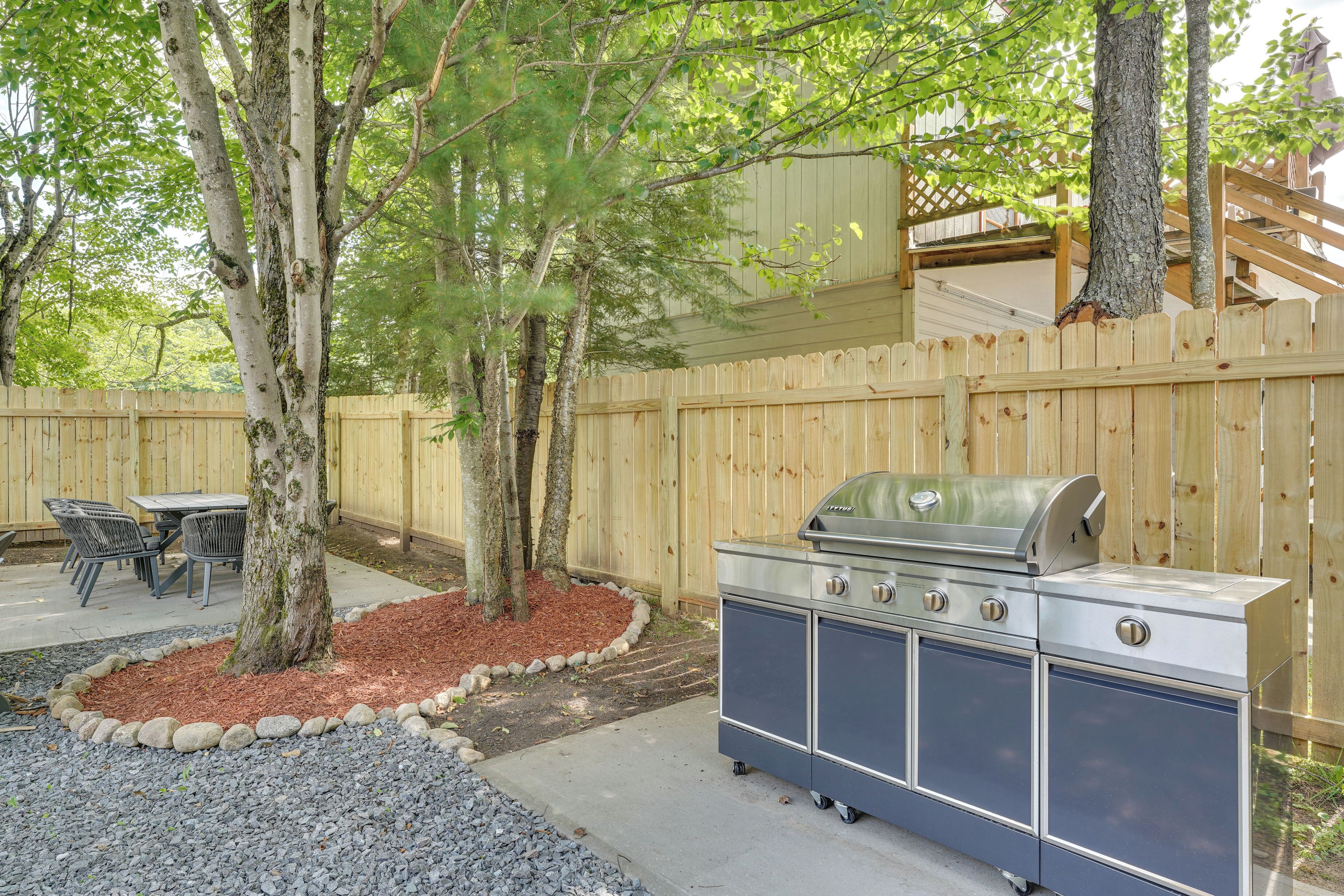 A backyard with a wooden fence, a barbecue grill, a table with chairs, and a tree.