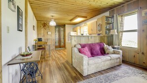 A cozy living room with a wooden ceiling, a sofa with a purple throw, a dining table with chairs, and a kitchen area with wooden cabinets and a window. - Scenic Mount Sopris View: Historic Alpine Cabin! (Glenwood Springs)