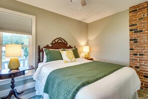 A bedroom with a bed, two bedside lamps, a window with blinds, a wooden nightstand, and a brick wall. - Updated Cotter Cottage: Walk to White River! (Cotter)