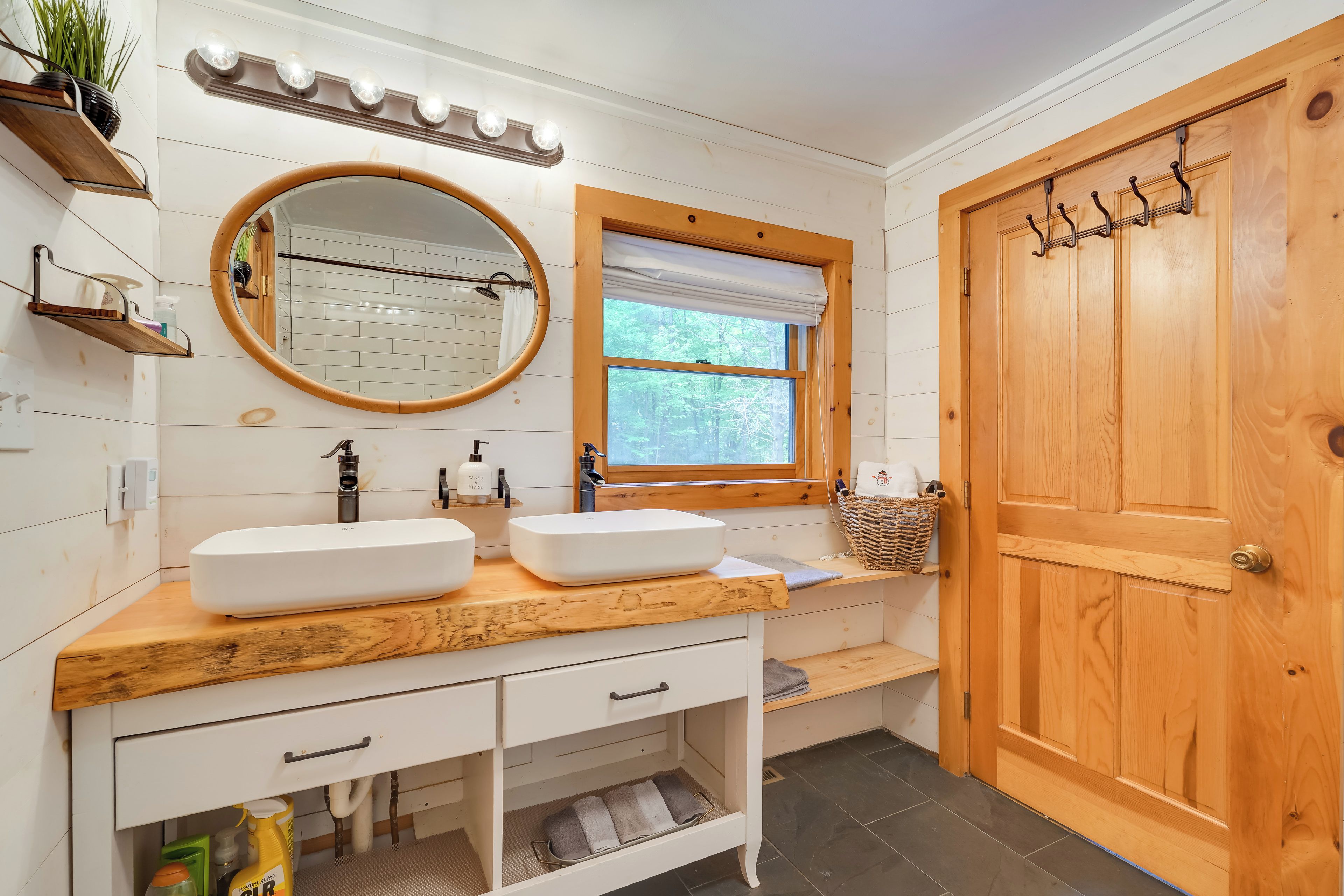 A bathroom with a wooden vanity, two sinks, a large mirror, a window with a view of trees, and a wooden door with hooks.