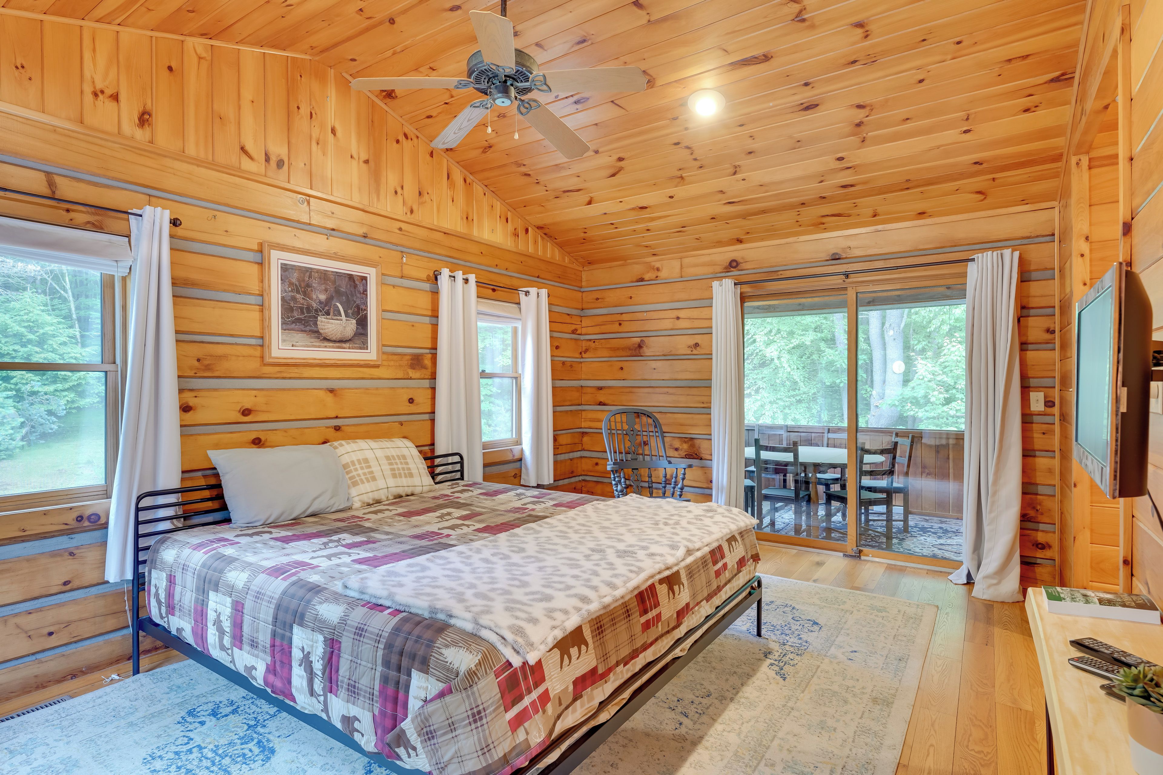 A wooden cabin bedroom with a bed, a ceiling fan, a dining table, and large windows.