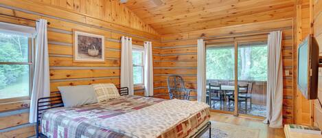 A wooden cabin bedroom with a bed, a ceiling fan, a dining table, and large windows.