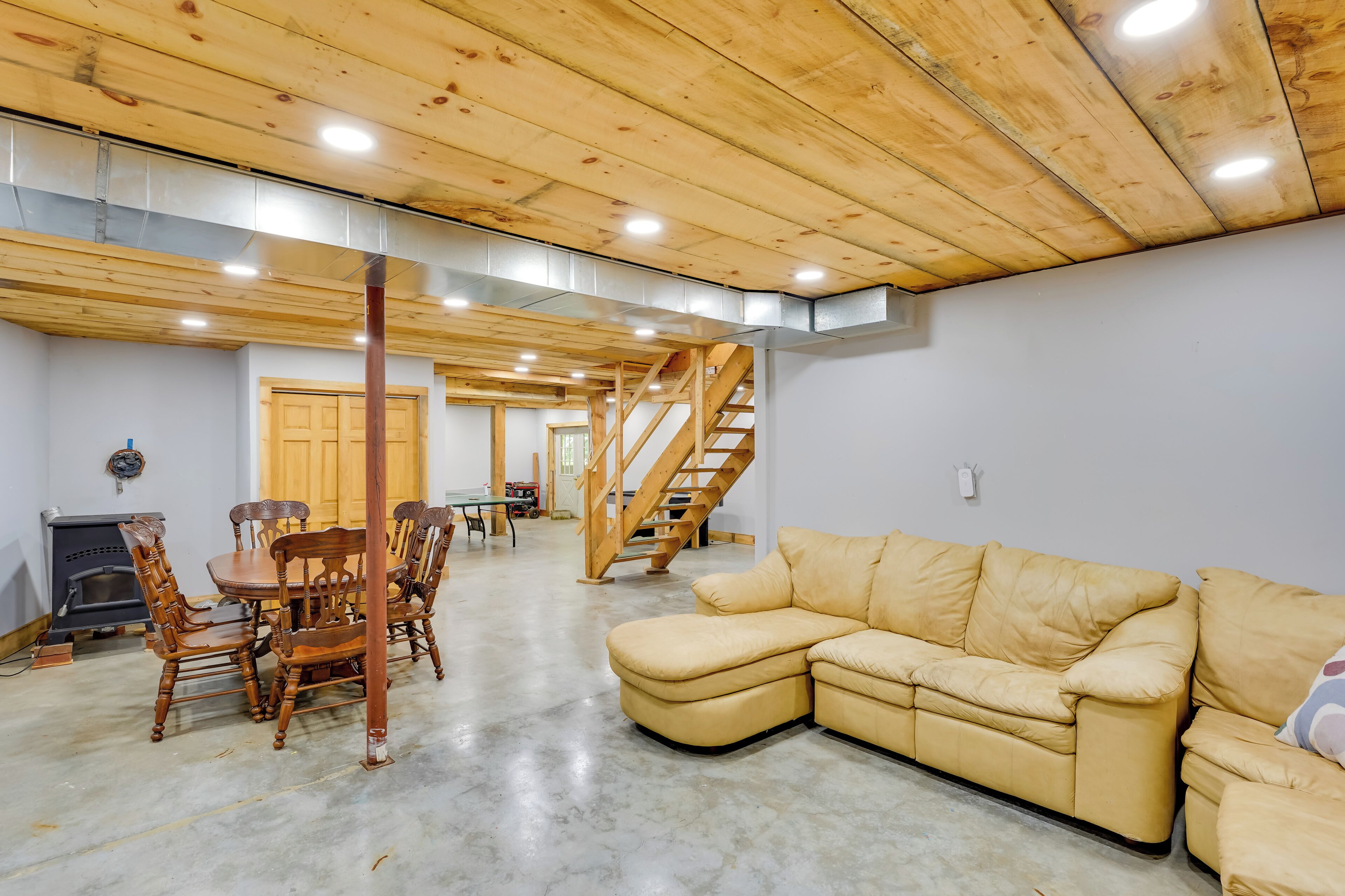 A spacious living area with a wooden ceiling, a beige sectional sofa, a dining table with chairs, and a staircase leading to an upper level.