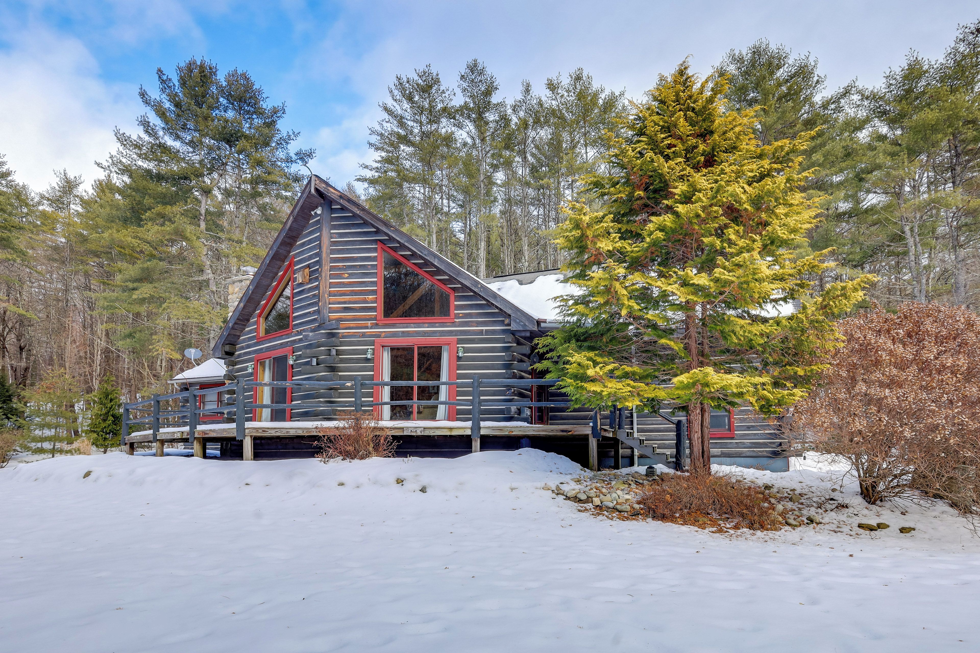 Une cabane en rondins de bois, avec un toit à forte pente, une fenêtre à cadre rouge et une véranda.