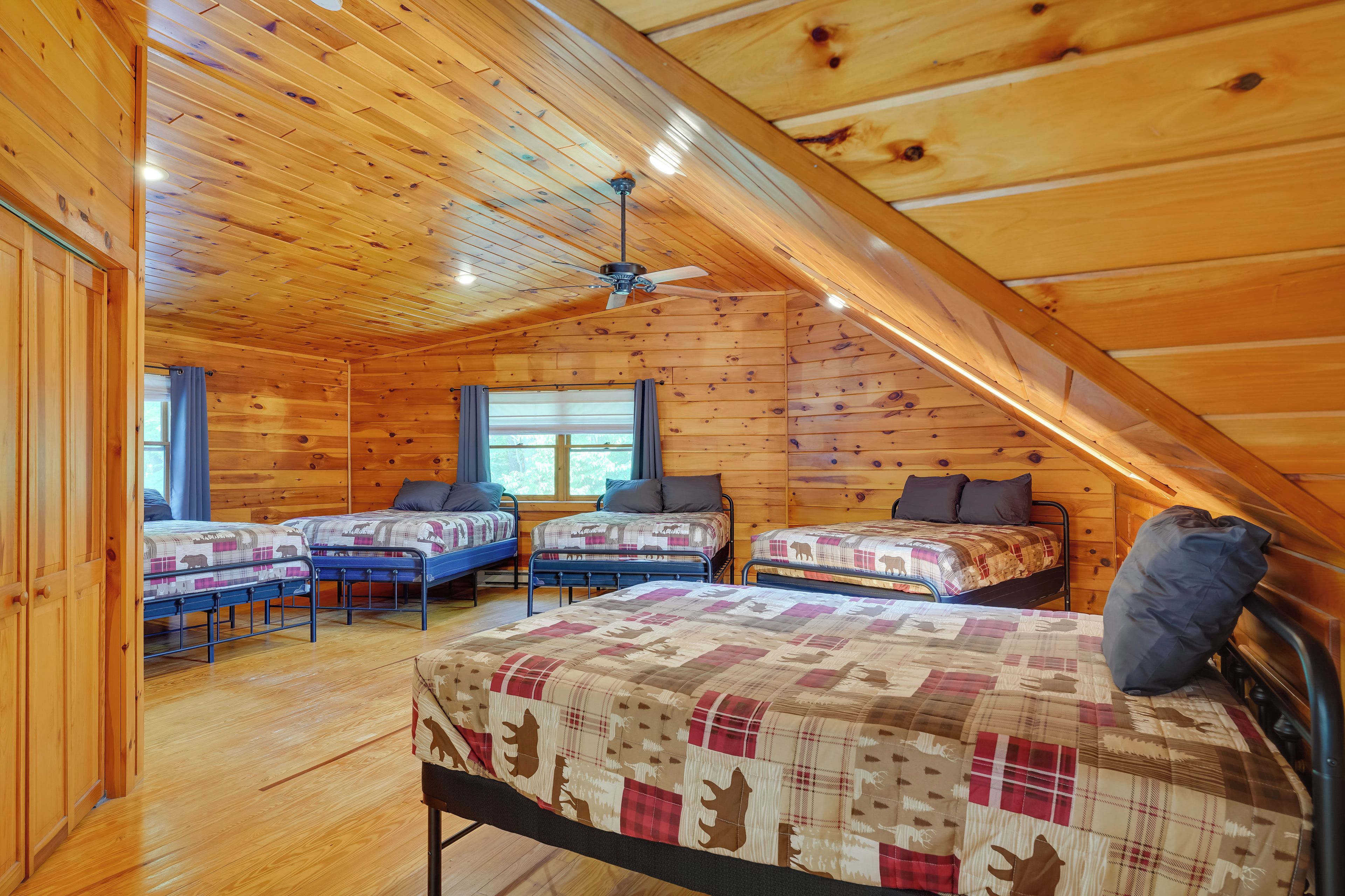 A cozy attic bedroom with wooden walls and a sloped ceiling. There are four beds arranged in a row, each with a patterned bedspread and pillows. A ceiling fan is installed in the center of the room.