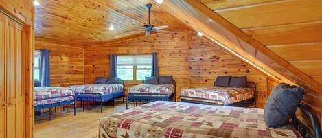 A cozy attic bedroom with wooden walls and a sloped ceiling. There are four beds arranged in a row, each with a patterned bedspread and pillows. A ceiling fan is installed in the center of the room.