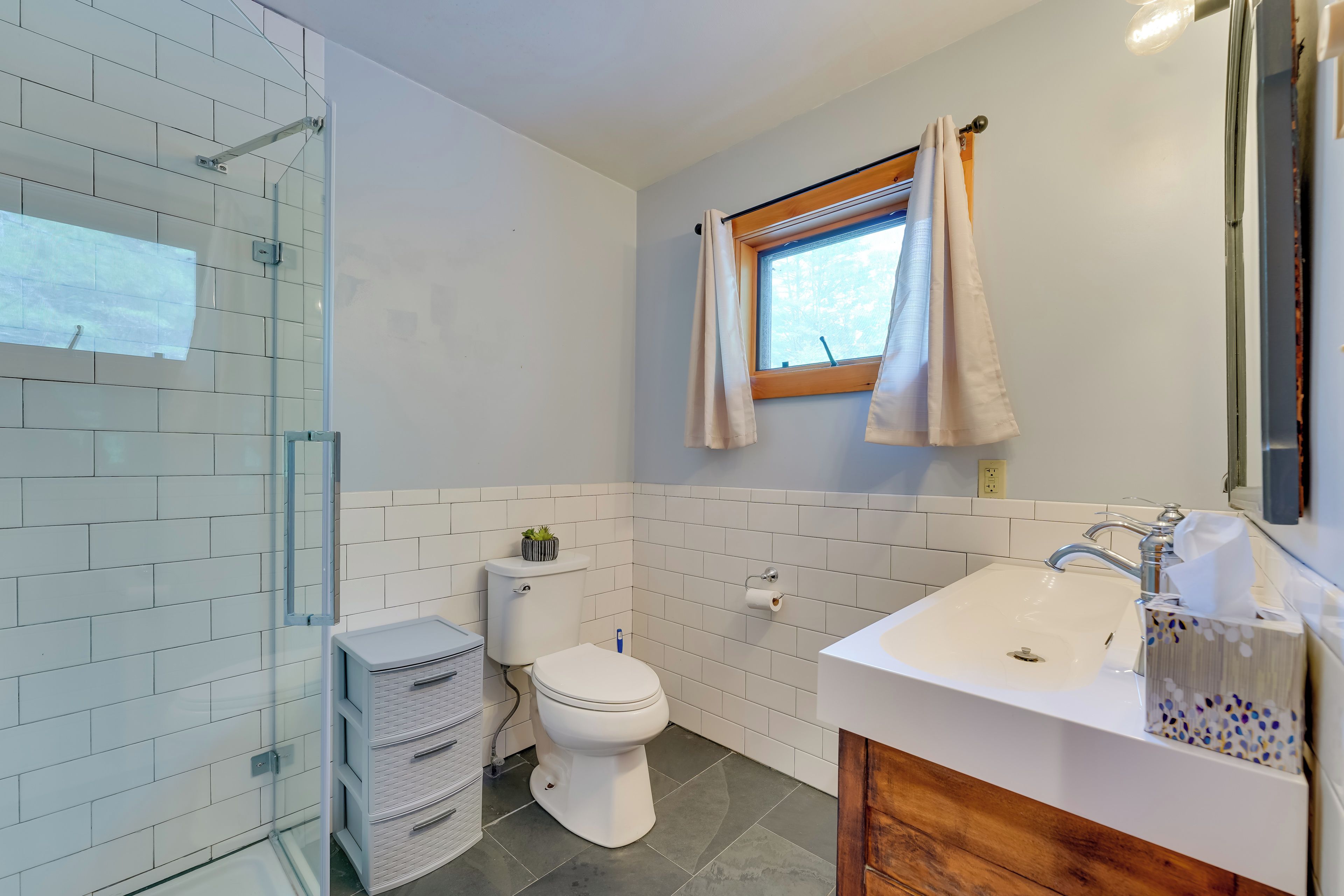 A modern bathroom with a glass shower enclosure, a toilet, a sink with a wooden vanity, and a window with curtains.
