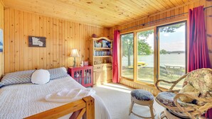 Een houten kamer met een bed, een klein tafeltje, een rieten stoel en een groot raam met uitzicht op de natuur.