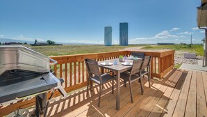 A wooden deck with a grill, dining table, and chairs, overlooking a grassy area and two tall buildings. - Bright Wyoming Retreat w/ Deck & Mountain Views! (Saratoga)