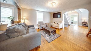 A spacious living room with a wooden floor, a grey sofa, a white armchair, a small coffee table, and a vintage trunk. There is a staircase leading to an upper level. - Historical Magnesia Springs Home, Near Kayaking! (Cambridge Springs)
