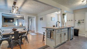 A modern kitchen with a central island, wooden dining table, and chairs. There is a built-in dishwasher and a hanging plant. - Historical Magnesia Springs Home, Near Kayaking! (Cambridge Springs)