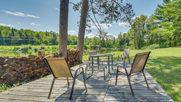 Una terraza de madera con mobiliario exterior, un muro de piedra y vistas a un lago rodeado de árboles.