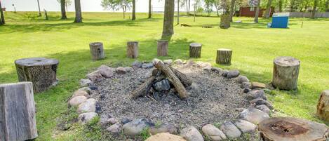 A fire pit surrounded by wooden stumps and rocks, set in a grassy area with trees and a building in the background.