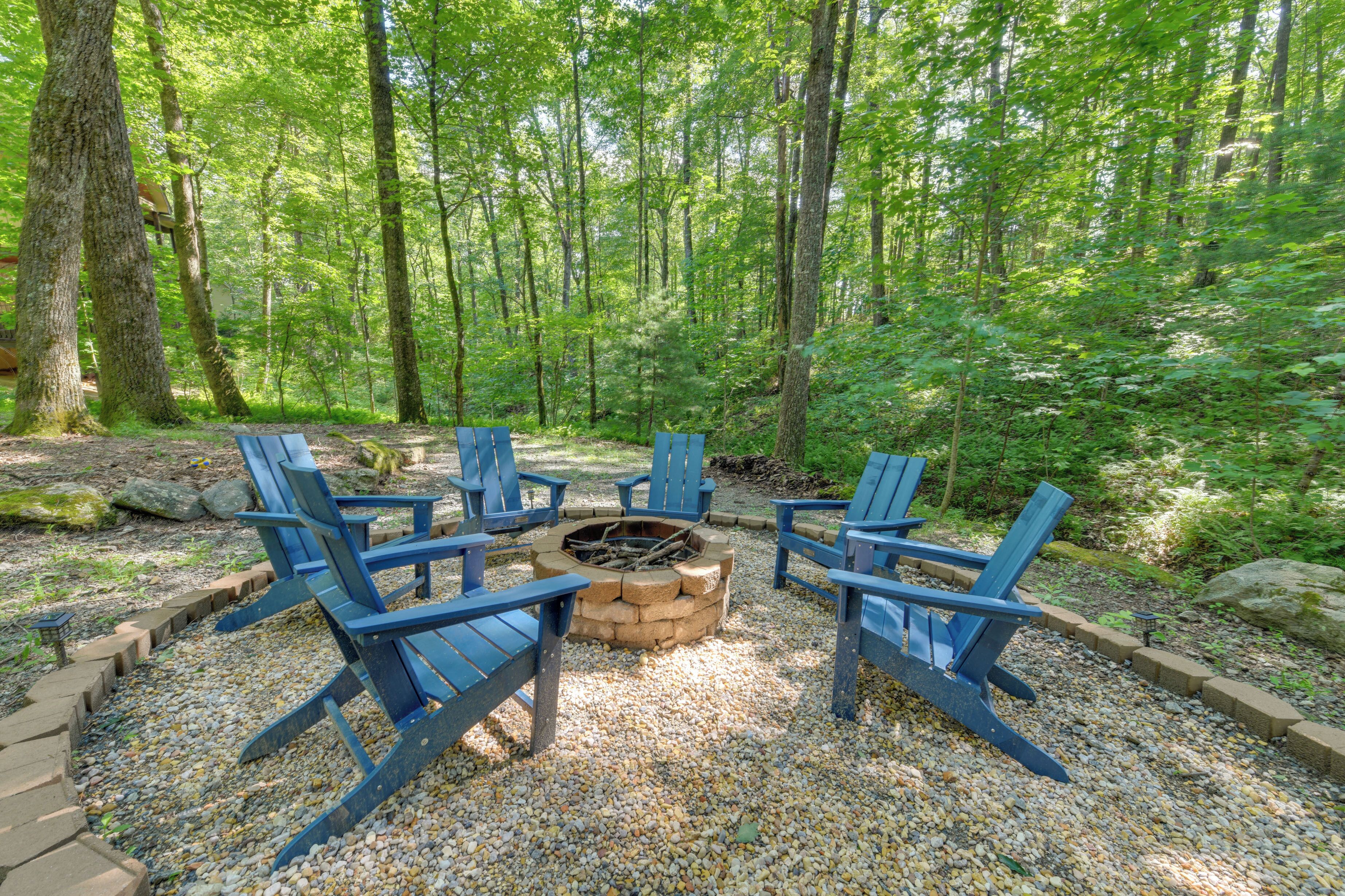A forested area with a fire pit and blue chairs arranged around it.