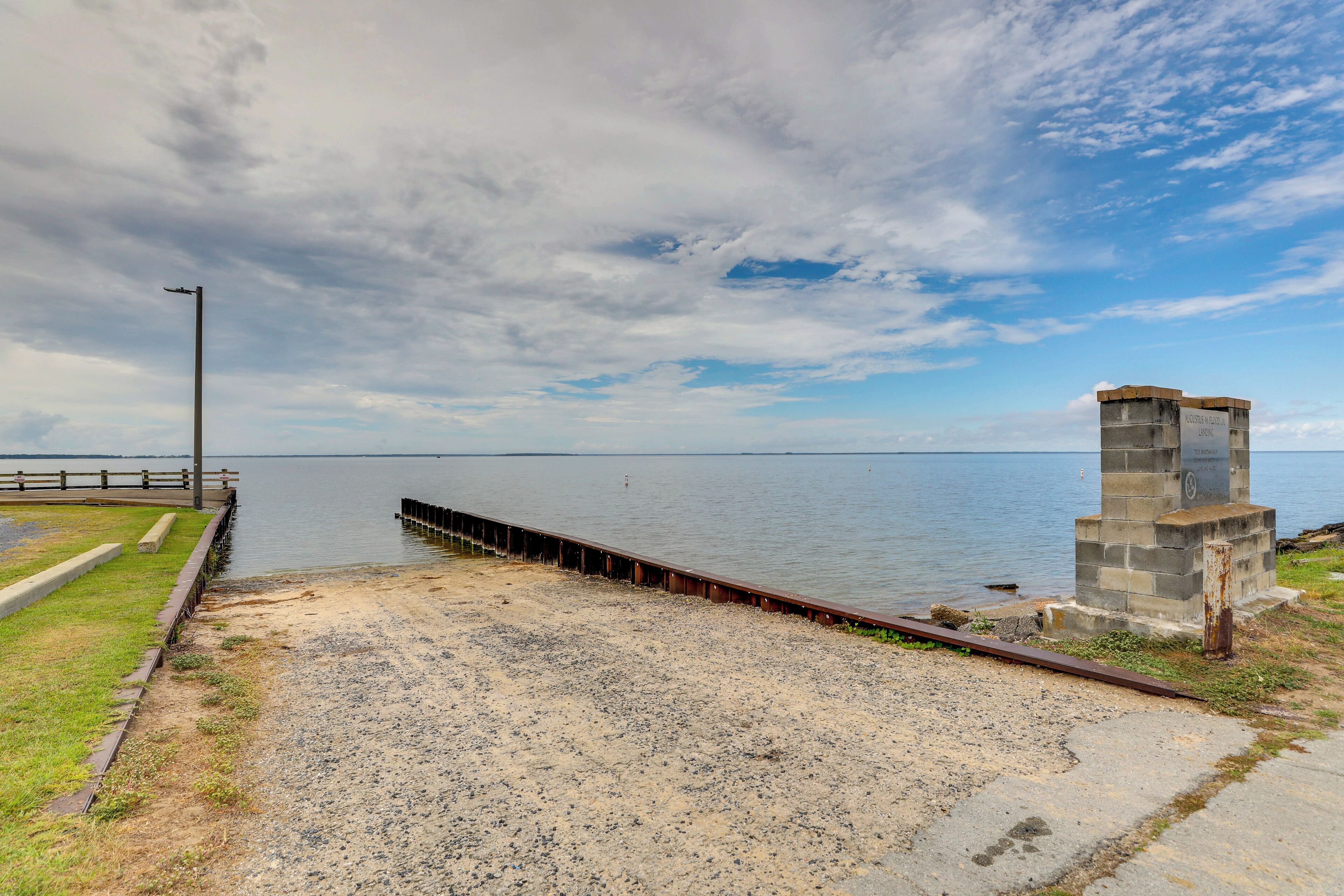 A waterfront area with a concrete pier extending into a calm body of water, a grassy area with a metal fence, and a stone monument.