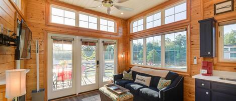 A cozy cabin interior with a wooden interior, a ceiling fan, a sofa with cushions, a coffee maker, and large windows.