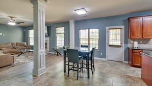 A spacious living room with a beige sofa, a glass coffee table, and a fireplace. There is a dining area with a dark wooden table and chairs. The kitchen has wooden cabinets and a countertop.