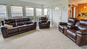 A living room with a brown leather sectional sofa, a single armchair, and a small table. There are large windows with a view of the outdoors. - Seiku Vacation Rental w/ Deck & Ocean Views (Sekiu)