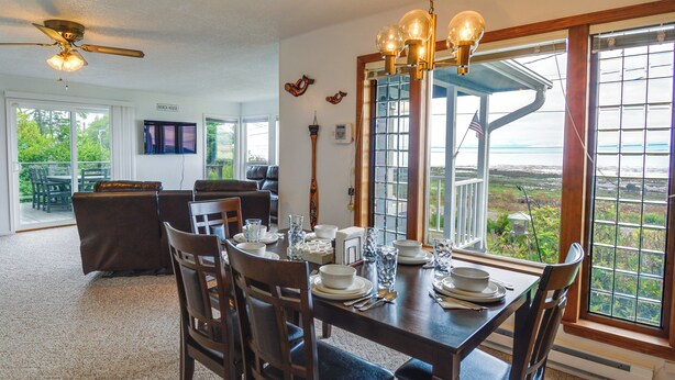 A dining area with a table set for four, surrounded by chairs. There is a television mounted on the wall and a view of the outdoors through a large window. - Seiku Vacation Rental w/ Deck & Ocean Views (Sekiu)
