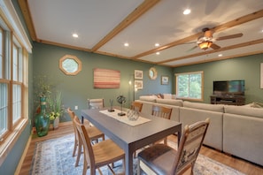 A dining area with a wooden table, chairs, and a vase with flowers. There is a sofa and a television in the background.