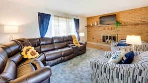 Ein Wohnzimmer mit einer Ledersofa, einem gemusterten Sessel, einem Kamin und einem darüber montierten Fernseher.