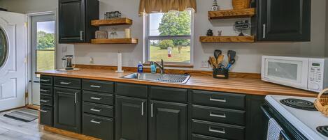 A kitchen with dark cabinets, a wooden countertop, a sink, a microwave, and a window with a view of greenery.