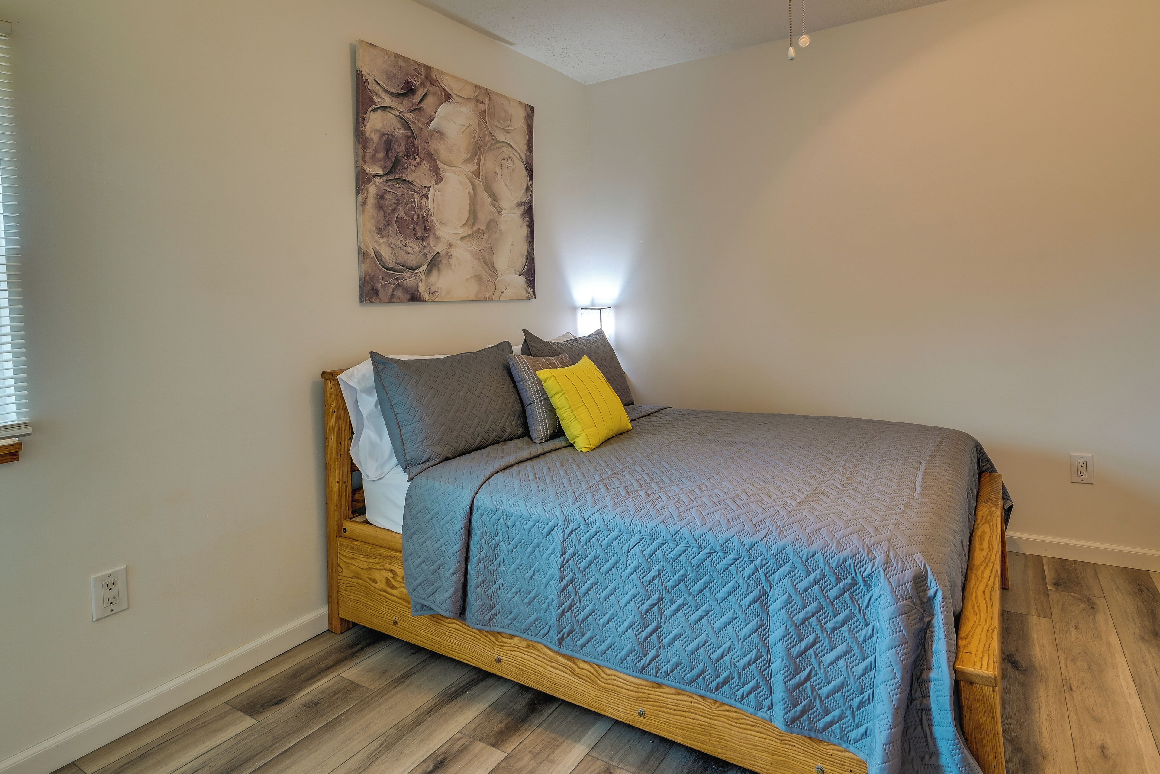 A bedroom with a wooden bed, a blue quilted bedspread, and a modern abstract painting on the wall.