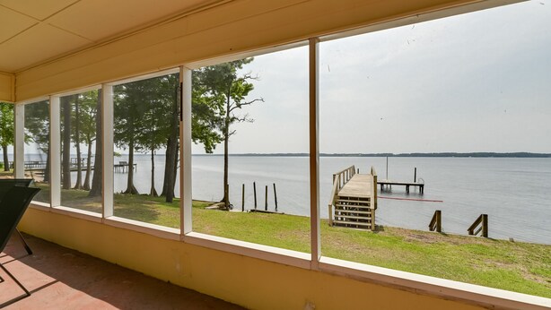 A covered outdoor area with large windows overlooking a lake, a wooden dock, and a grassy area. - Riverfront Edenton Condo w/ Porch & Water Views! (Edenton)