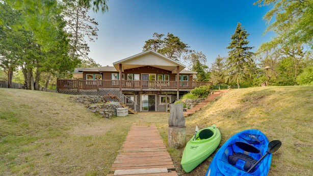 A wooden pathway leading to a two-story house with a porch, surrounded by trees and a kayak on the right. - Lakefront Wisconsin Home - Deck, Fire Pit & Kayaks (Stone Lake)