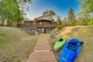 A wooden pathway leading to a two-story house with a porch, surrounded by trees and a kayak on the right.