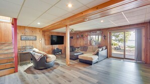 A spacious living room with wooden floors, a large flat-screen TV, and a sliding glass door leading to an outdoor area. - Lakefront Wisconsin Home - Deck, Fire Pit & Kayaks (Stone Lake)