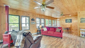A cozy cabin interior with a red sofa, a leather chair, a red wood stove, and a dining area with a wooden table and chairs. - Lakefront Wisconsin Home - Deck, Fire Pit & Kayaks (Stone Lake)