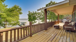 A wooden deck with a railing overlooking a body of water, a covered seating area with cushions, and a view of trees and a distant shoreline. - Lakefront Wisconsin Home - Deck, Fire Pit & Kayaks (Stone Lake)