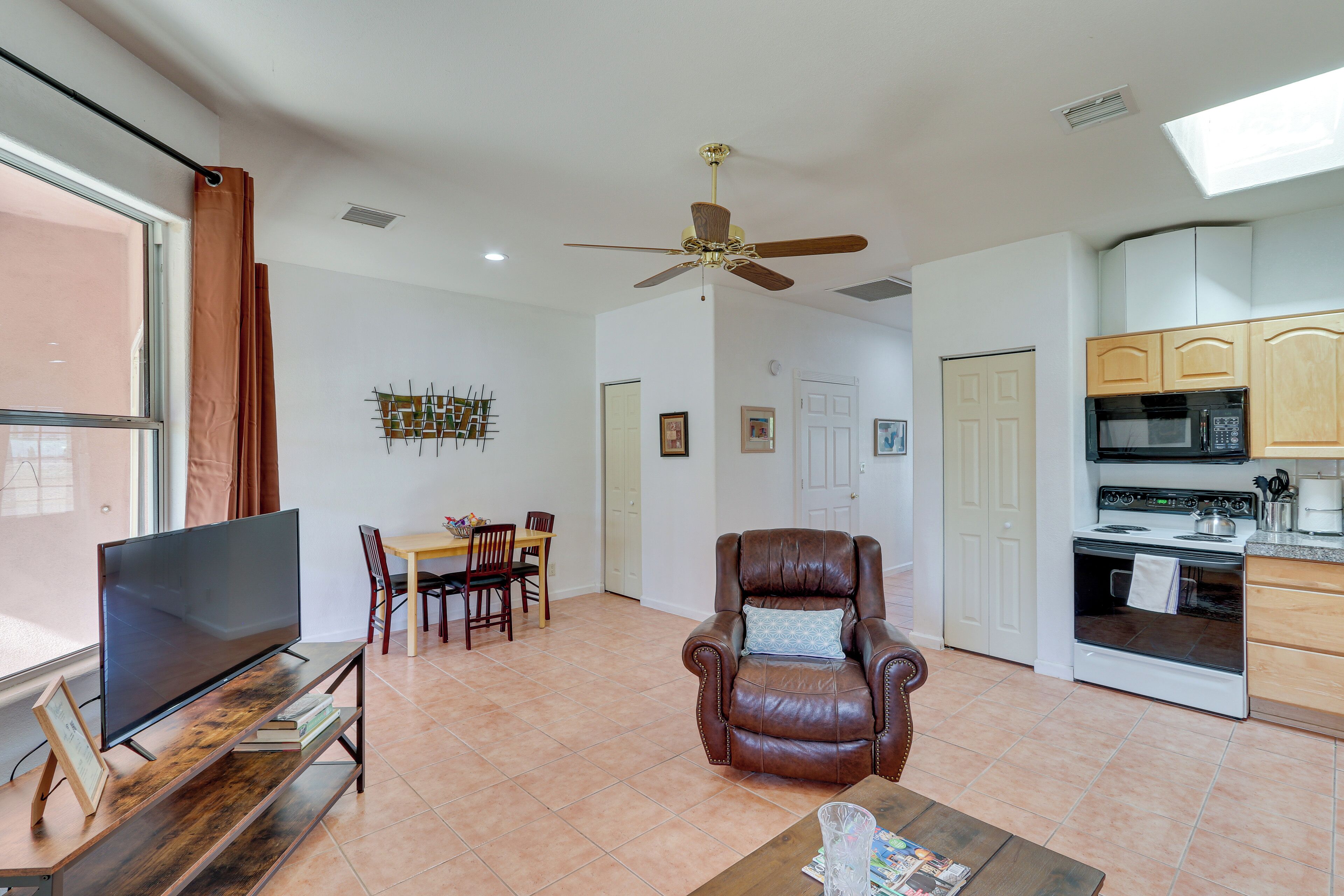 A modern living room with a flat-screen TV, a leather armchair, a dining table with chairs, and a kitchen area with a microwave and stove.