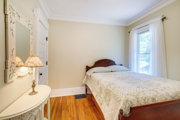 A bedroom with a wooden bed, a white dresser, a mirror with floral designs, and a window with white curtains. - Cozy Michigan Retreat w/ Deck & Gas Grill! (Frankfort)