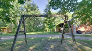 A wooden swing set in a grassy yard with trees and a wooden structure in the background. - Minnesota Cabin Rental: 3 Mi to Pelican Lake! (Pequot Lakes)