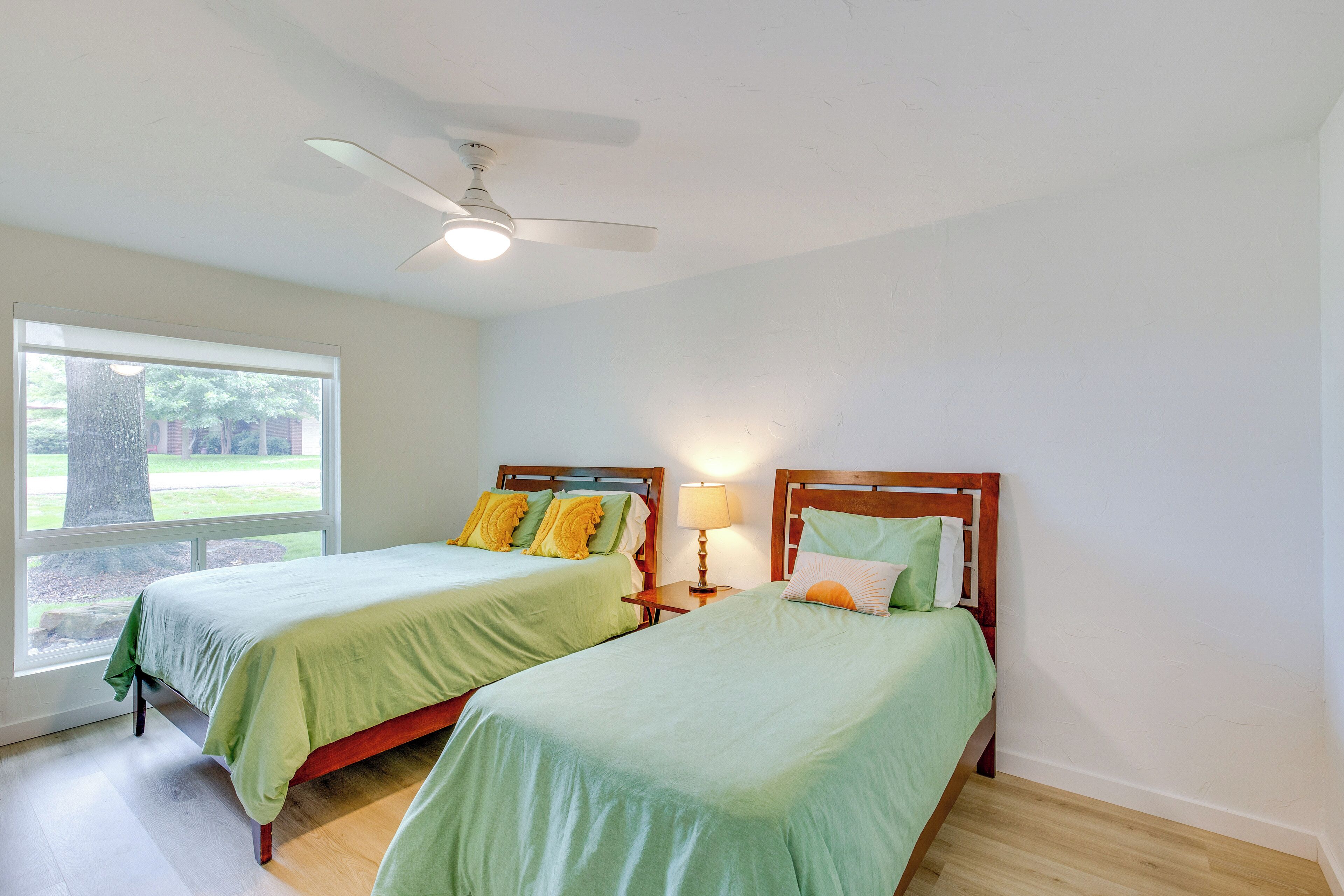 A bedroom with two beds, a ceiling fan, a window with a view of greenery, and a nightstand with a lamp.