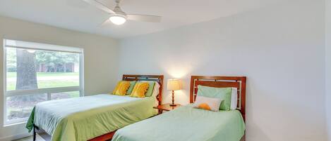 A bedroom with two beds, a ceiling fan, a window with a view of greenery, and a nightstand with a lamp.