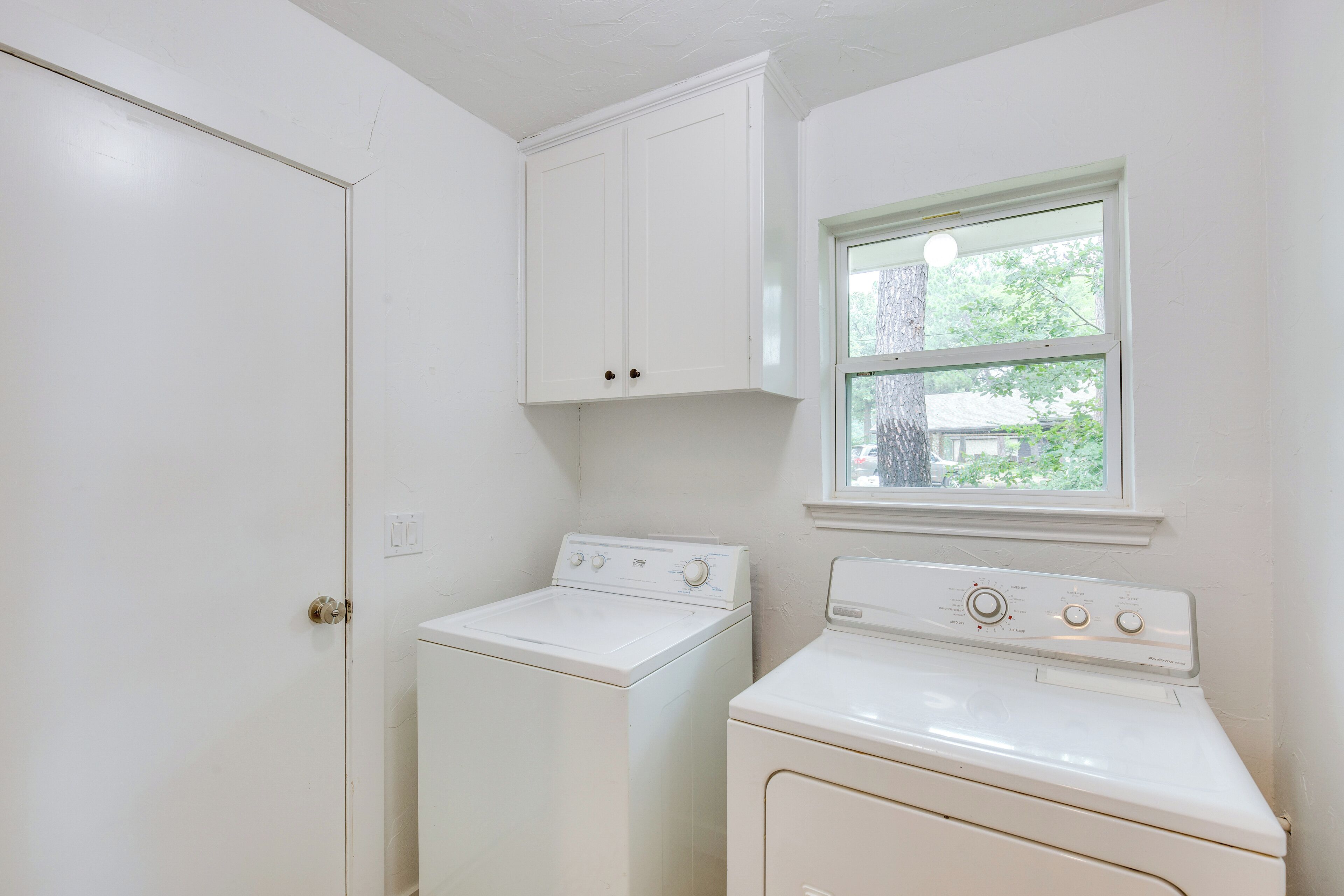 A laundry room with a washing machine and dryer, a window with a view of trees, and a white door.
