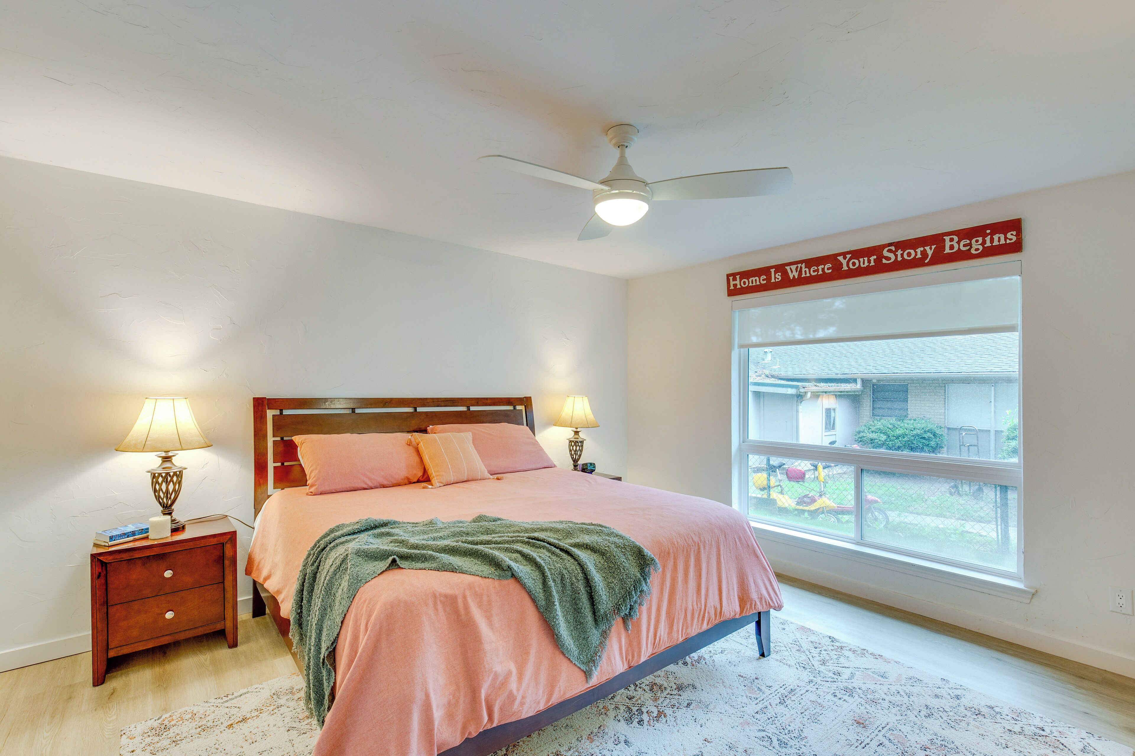 A bedroom with a bed, two bedside tables with lamps, a ceiling fan, a large window, and a motivational quote on the wall.