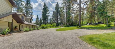 A residential property with a gravel driveway, a house with a brown roof, and a forested area with tall trees.