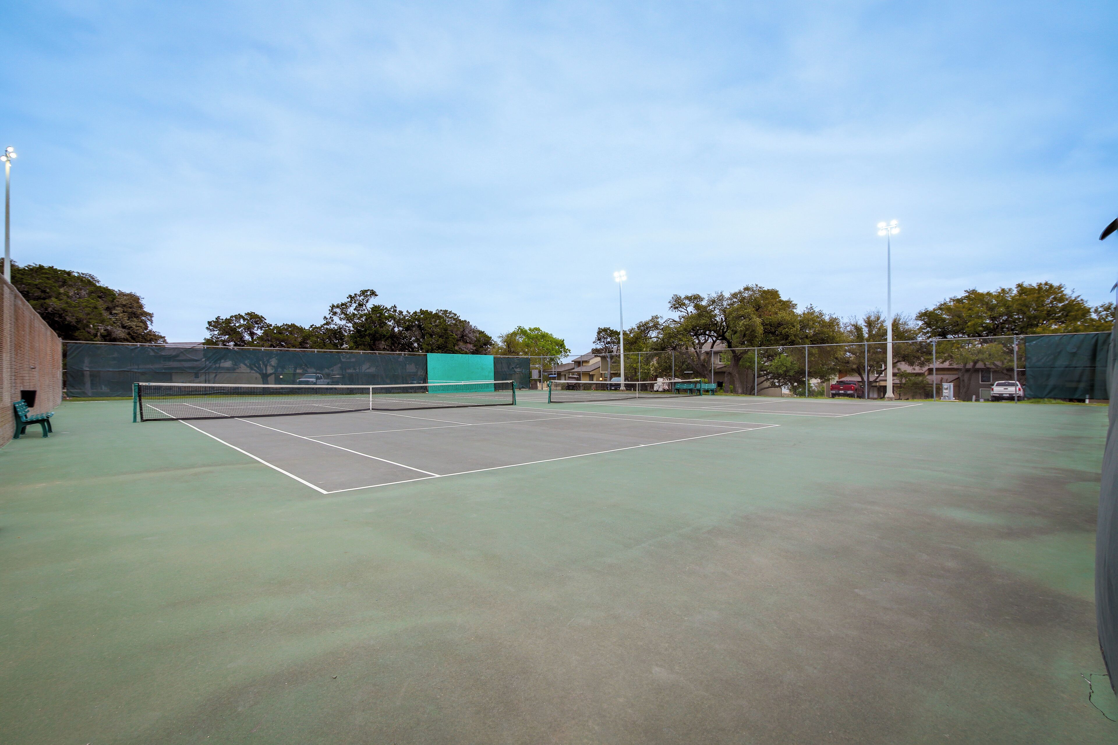An outdoor tennis court with a net, surrounded by a fence, and trees in the background.