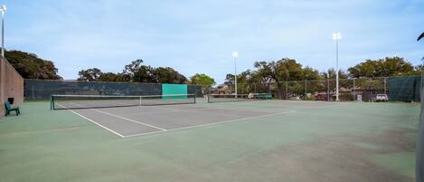 An outdoor tennis court with a net, surrounded by a fence, and trees in the background.