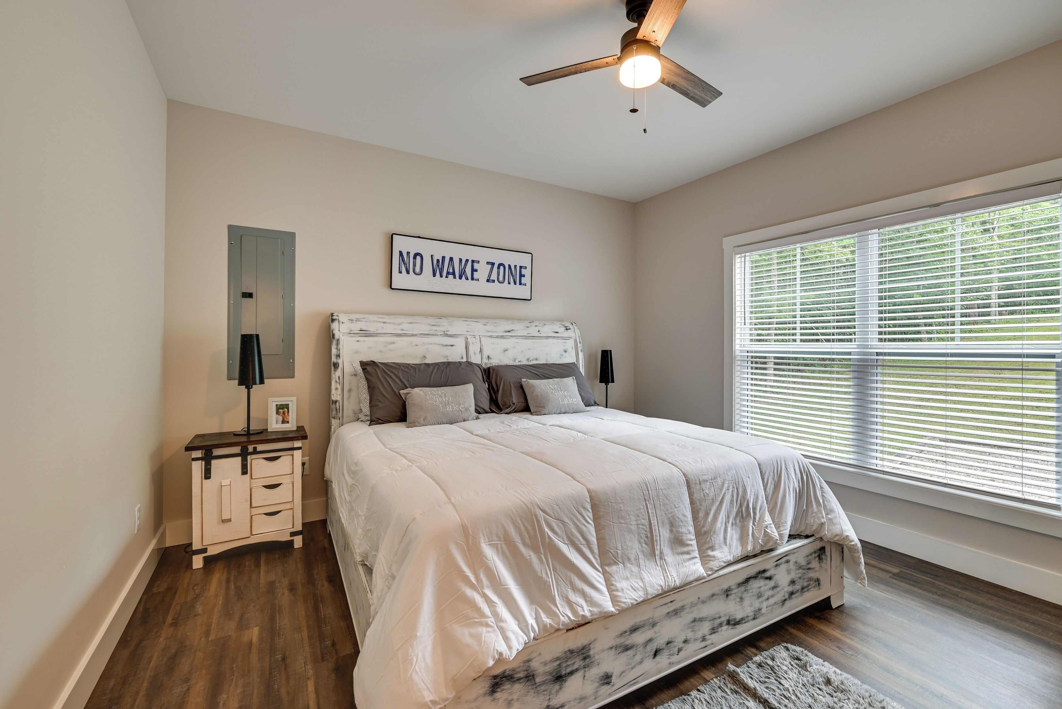 A bedroom with a bed, a nightstand, a mirror, a ceiling fan, and a window with blinds.