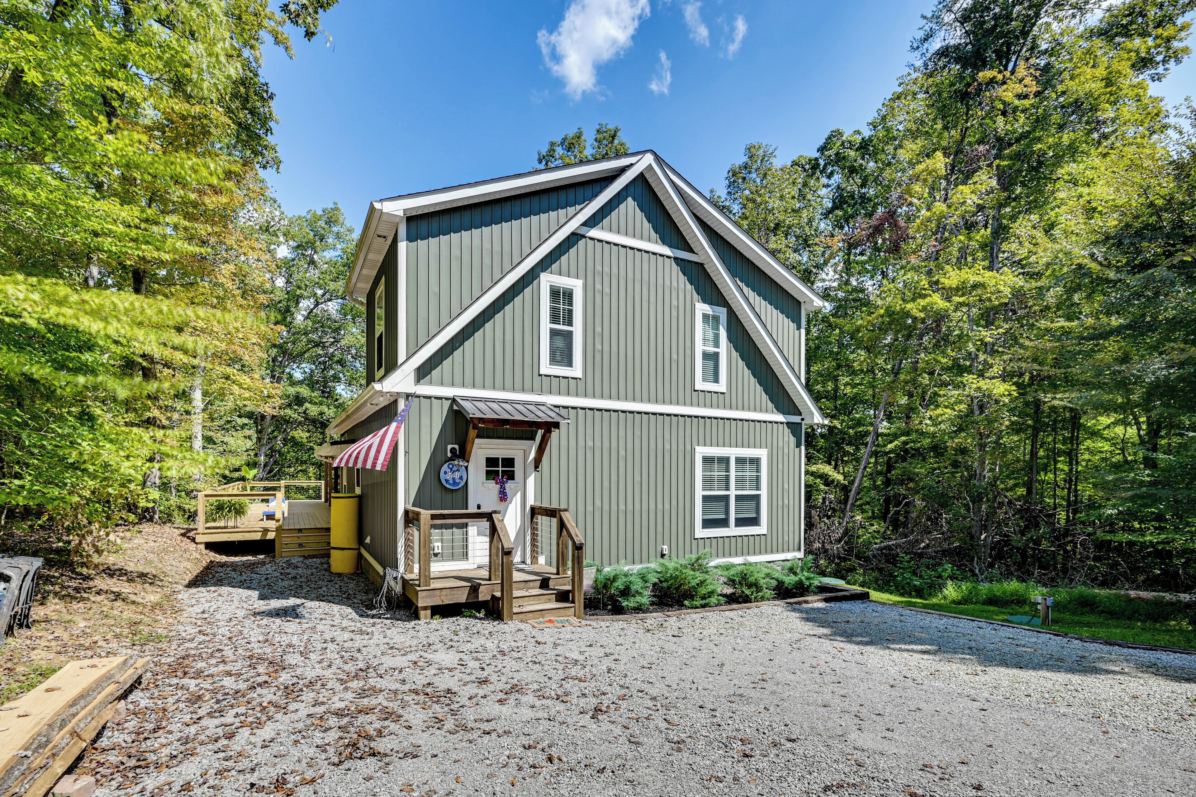 A two-story house with a front porch and a small deck, surrounded by trees and a gravel driveway.