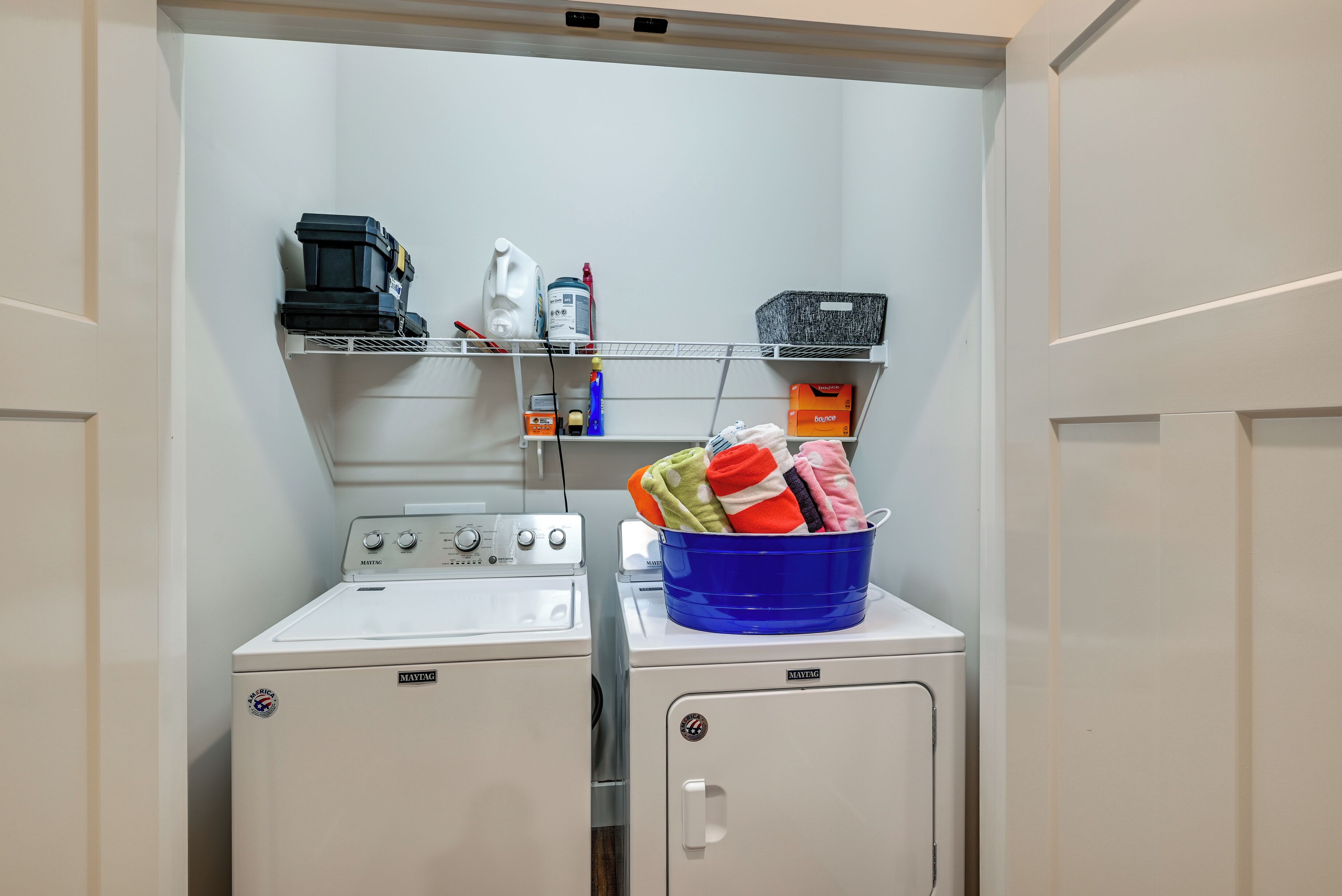 Un área de lavandería con dos lavadoras Maytag, un estante con productos de limpieza y una cesta azul con ropa.